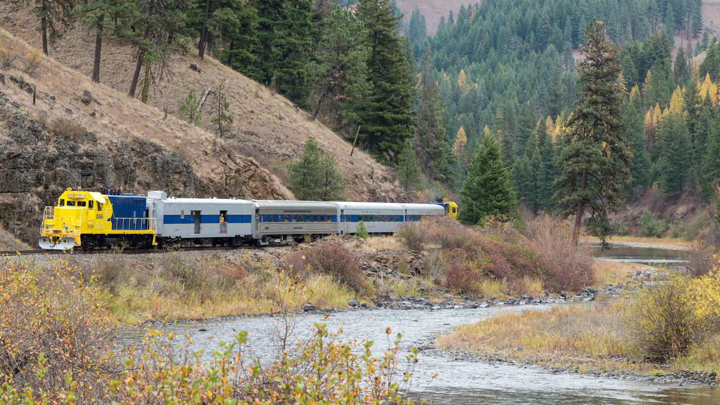 A passenger train passes along a large creek in the Eagle Cap Wilderness.