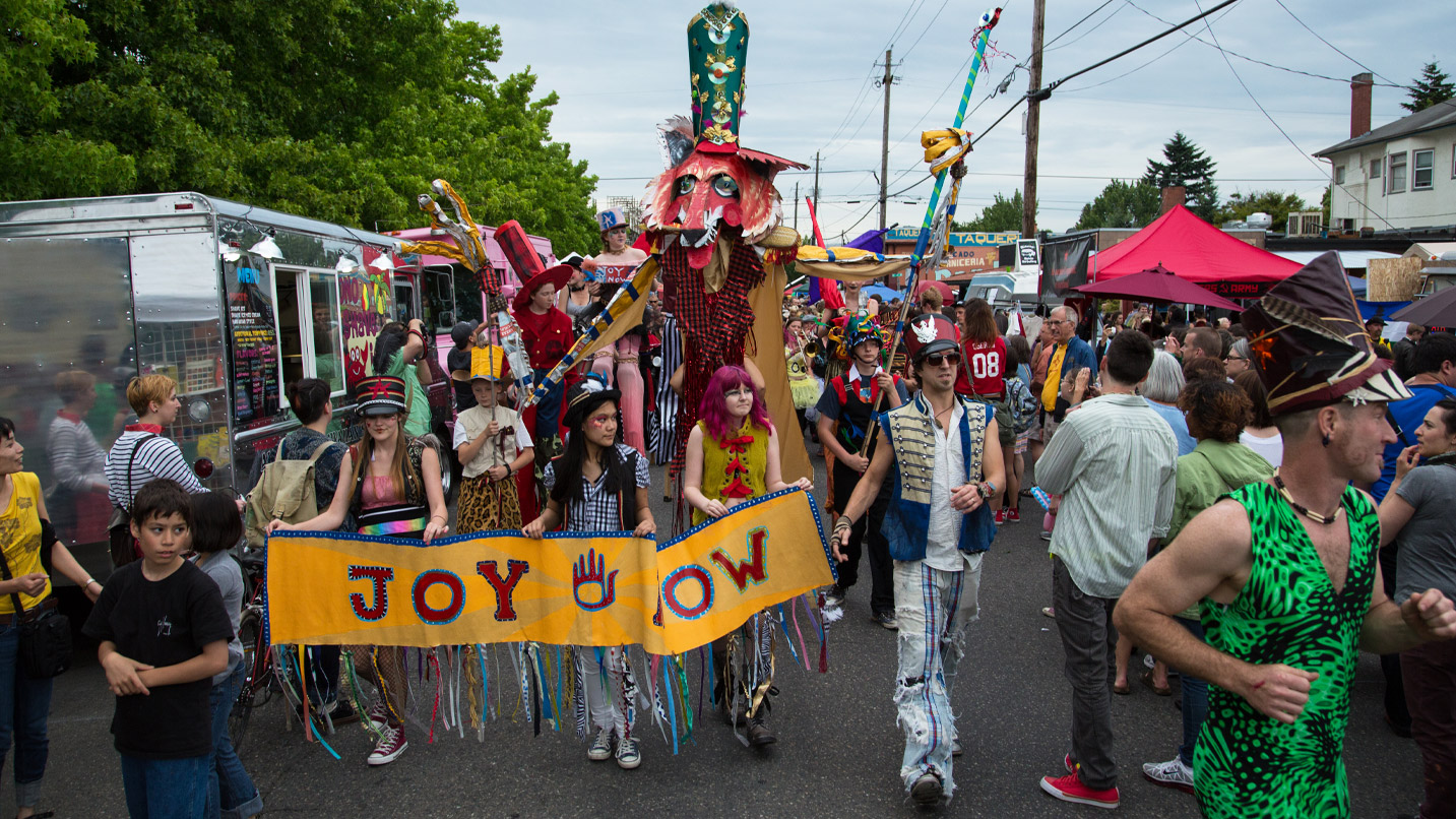 Several people dressed up in colorful clothing and accessories at a street fair. Three women hold a banner and a giant cardboard wolf sculpture is behind them.