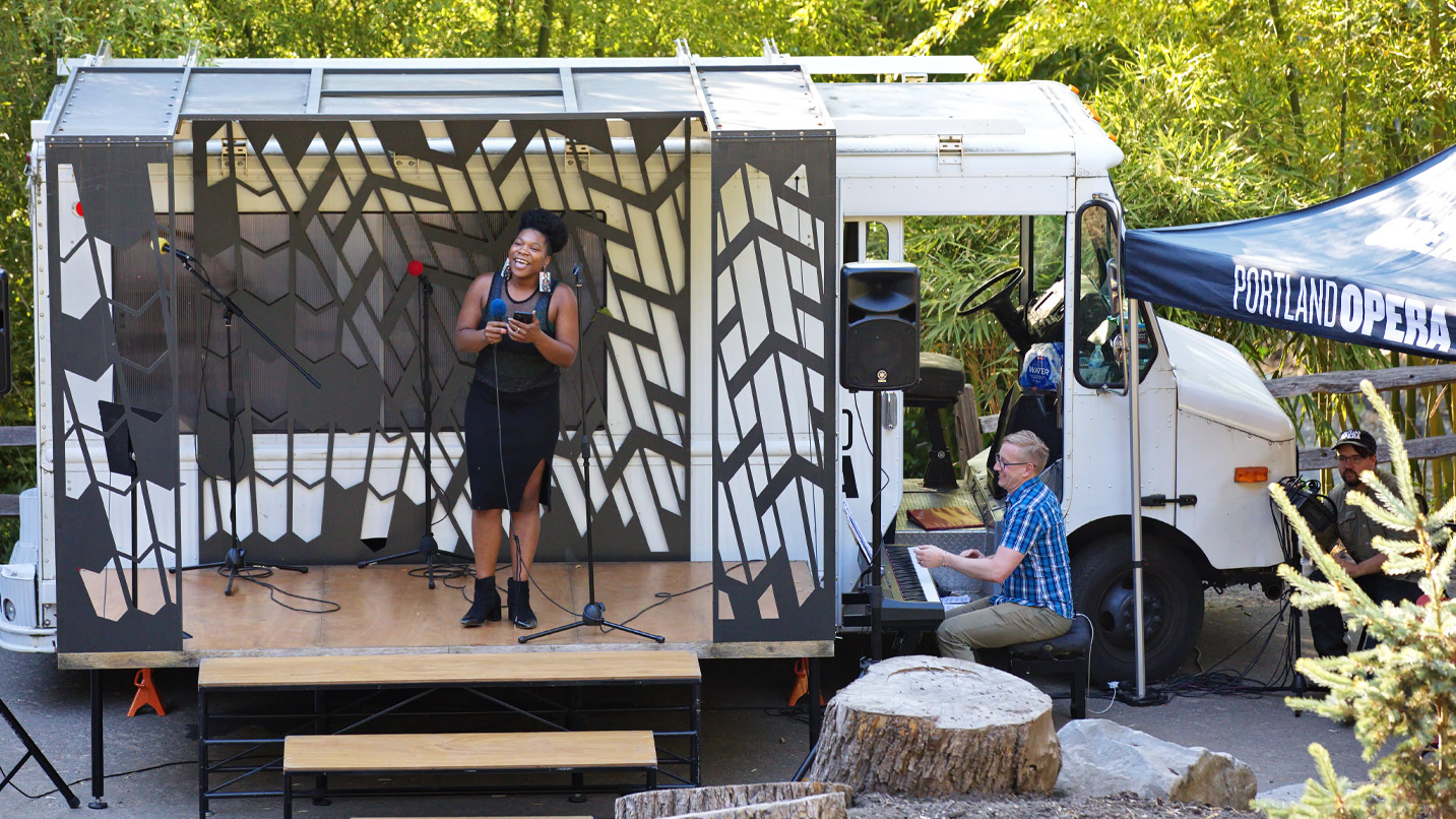 A woman sings opera from a food truck with a makeshift stage. A man plays a keyboard nearby.