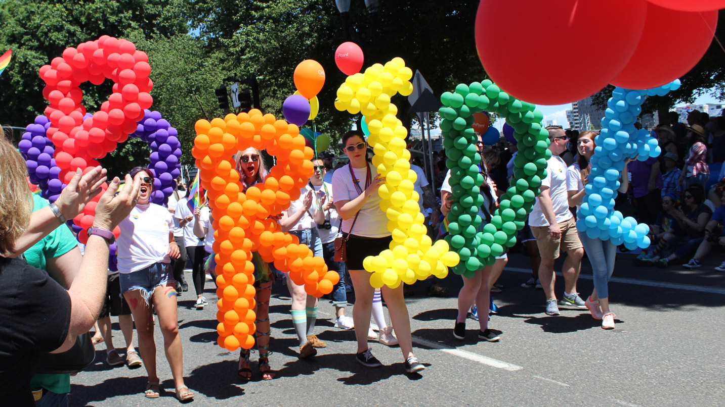 Several people in a parade holding letters built of different colored balloons spelling out PRIDE.