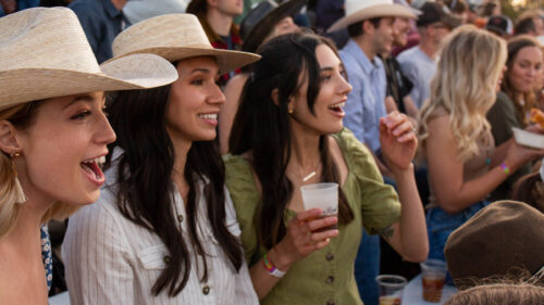 A lively crowd, with women in cowboy hats holding drinks, cheers and enjoys an outdoor event.