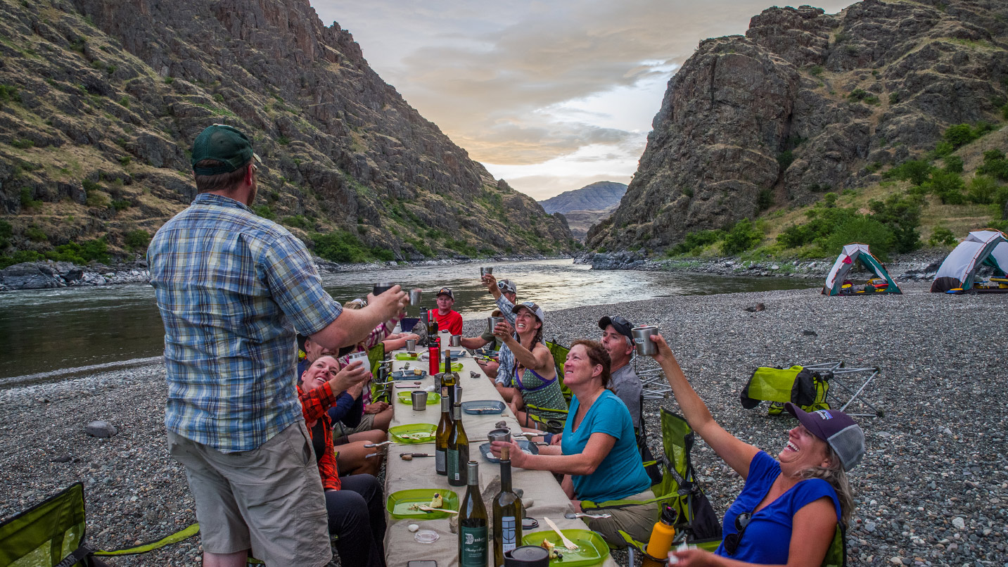 People sit at a long dining table on a rocky riverbank, raising drinks in a toast. Camping tents are set up nearby, surrounded by steep, rocky hills under a cloudy sky.