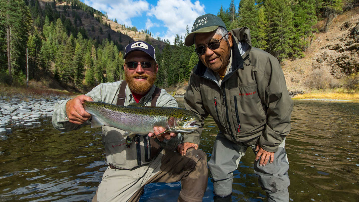 Two people hold a large fish together, smiling in shallow river water surrounded by forested mountains. One man wears a "FLY SHOP" cap and a jacket labeled "REDINGTON."