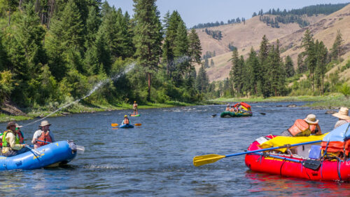 People in colorful rafts paddle on a wide river surrounded by lush forests and distant hills, under a clear blue sky.