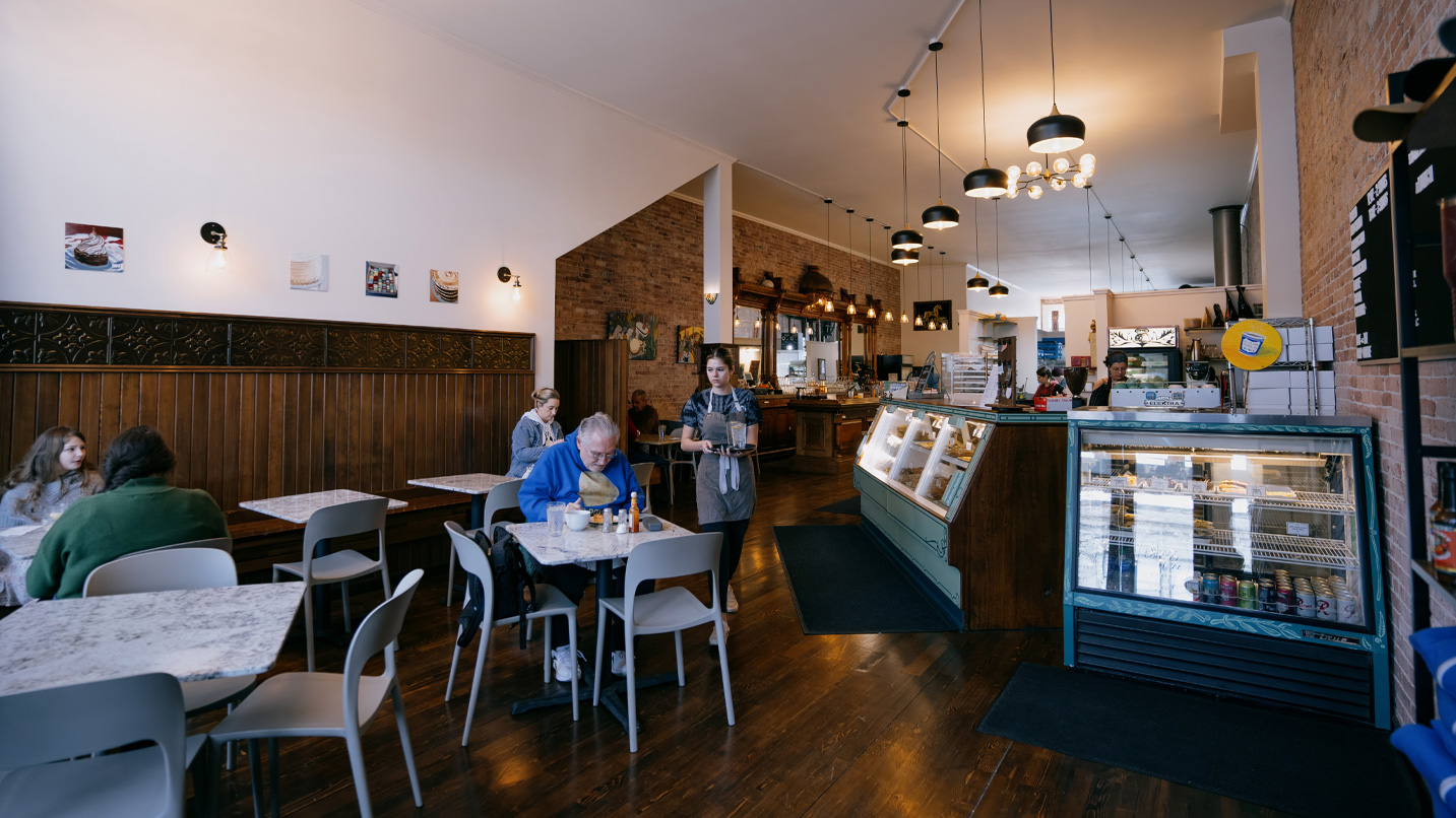 A café features people seated at tables, eating and conversing. A server carries a tray past refrigerated displays. Warm lighting and decor create a cozy atmosphere.