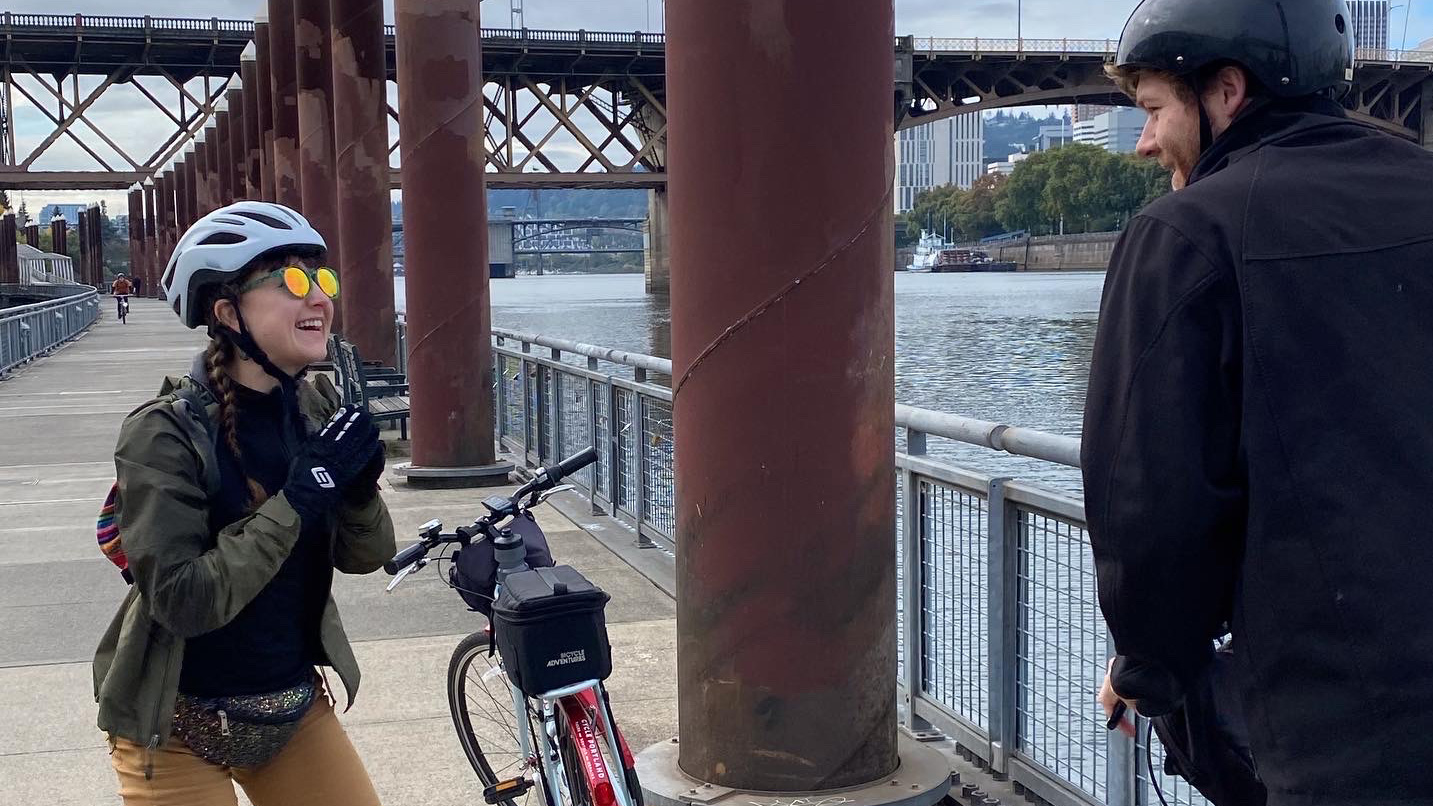 Two cyclists, wearing helmets, converse and laugh near their parked bikes on a riverside path. A rusted bridge and cityscape form the backdrop, with water and buildings visible.