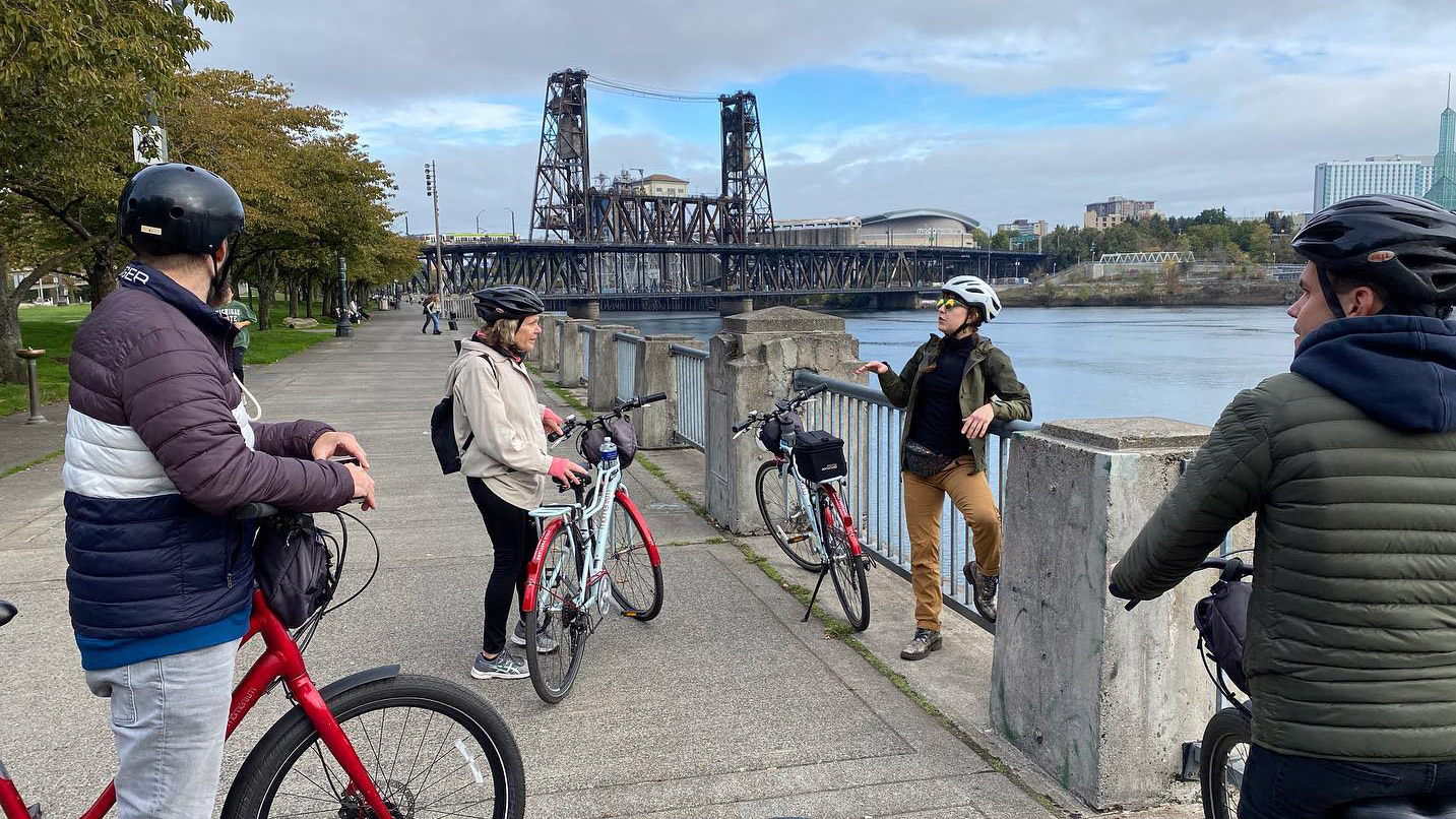 Cyclists gather with their bikes along a riverside path. One person energetically talks while others listen. Behind them, a steel bridge spans the calm river under a partly cloudy sky.