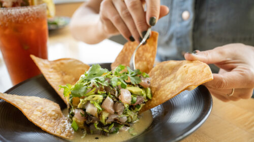 A person holds a crispy tortilla chip while using a fork to eat ceviche, garnished with cilantro, in a black bowl. A red drink with a salted rim is nearby.