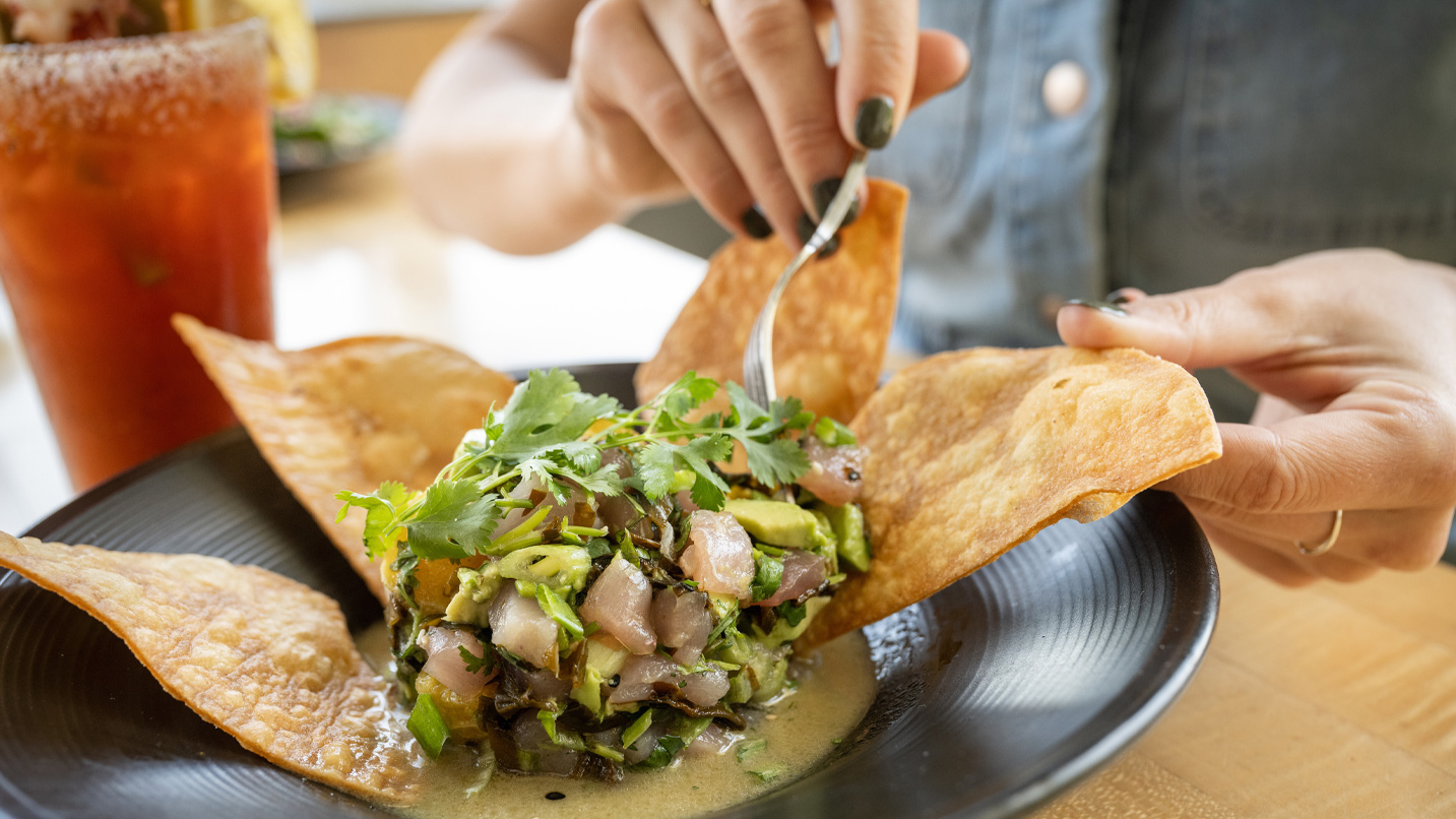 A person holds a crispy tortilla chip while using a fork to eat ceviche, garnished with cilantro, in a black bowl. A red drink with a salted rim is nearby.