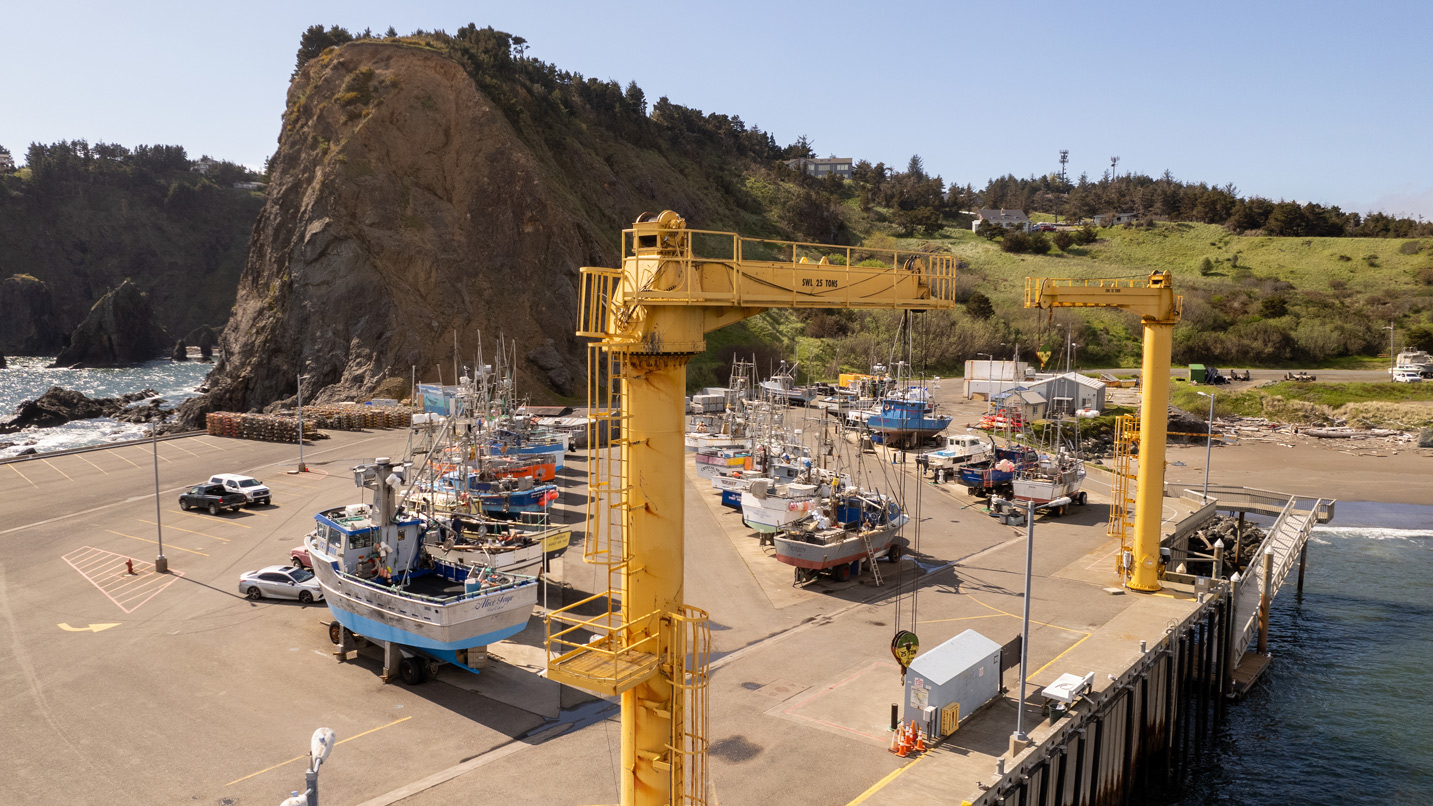 Boats are parked on a dock, under two large yellow cranes labeled "SWL 25 TONS," near a rocky coastline with grassy hills and a few parked vehicles nearby.