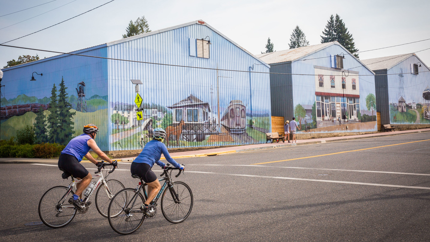 Two cyclists ride past mural-painted farm buildings.