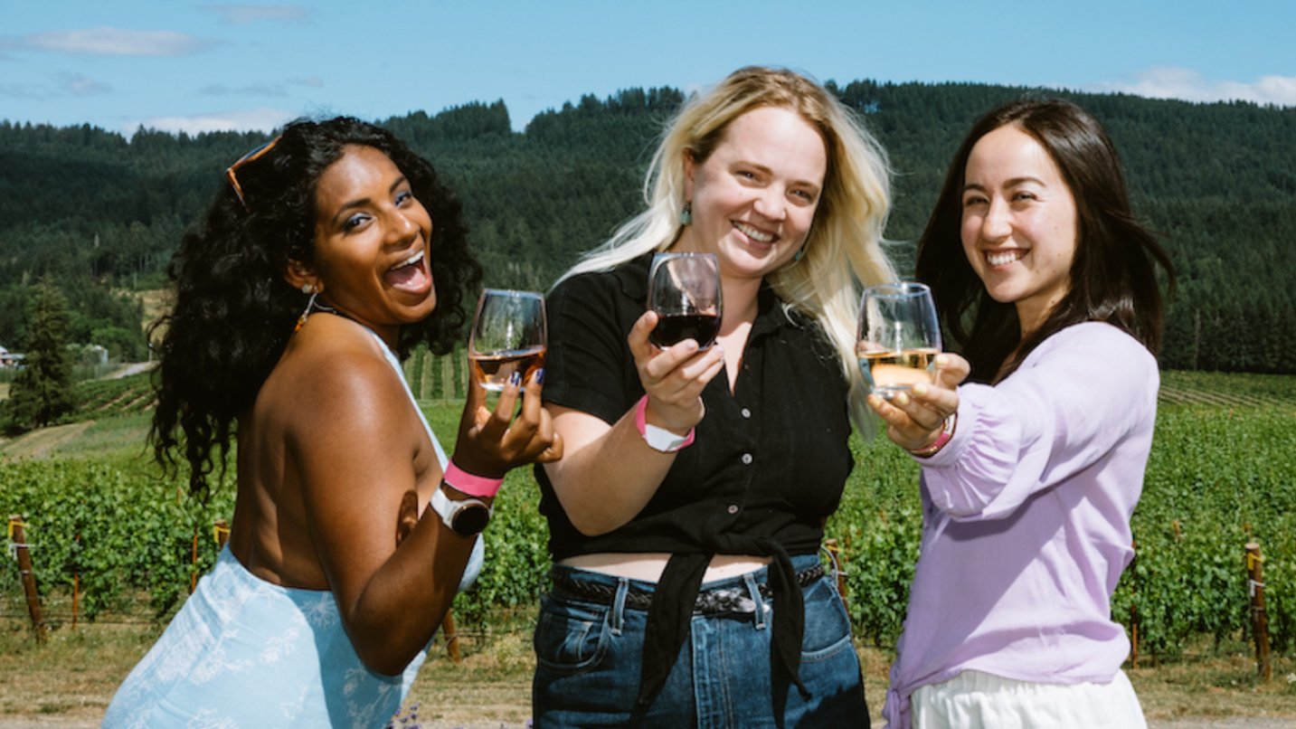Three women hold glasses of wine and smile in front of a vineyard