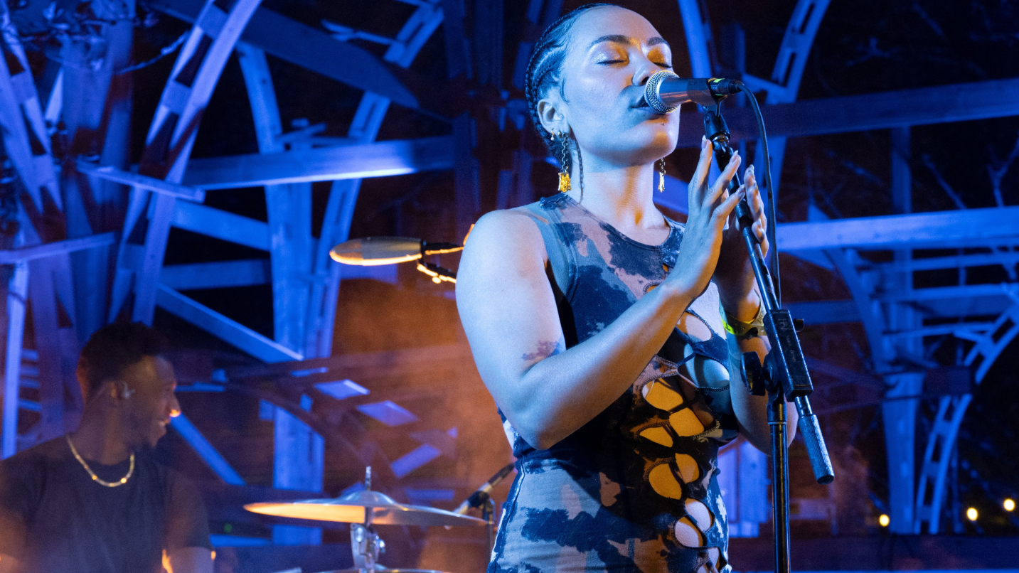 Woman sings into microphone against dark sky and purple lights in background