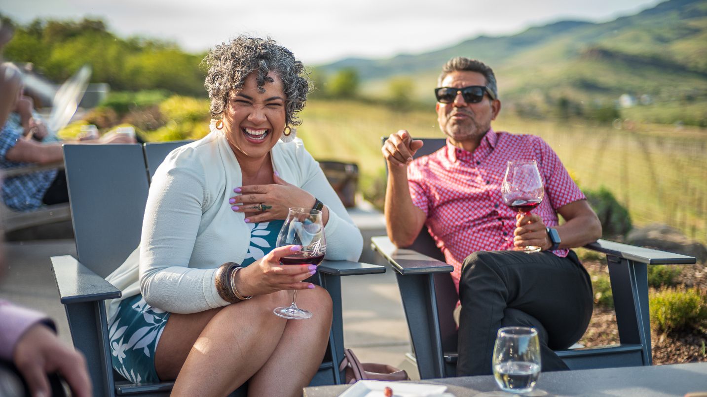 Two people sit on chairs holding wine glasses with mountains blurred in background