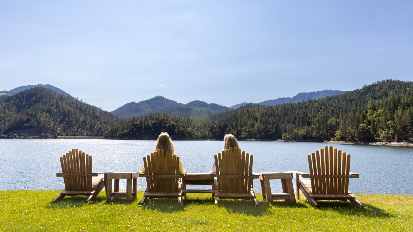 Two people sit in Adirondack chairs next to a lake surrounded by hills.