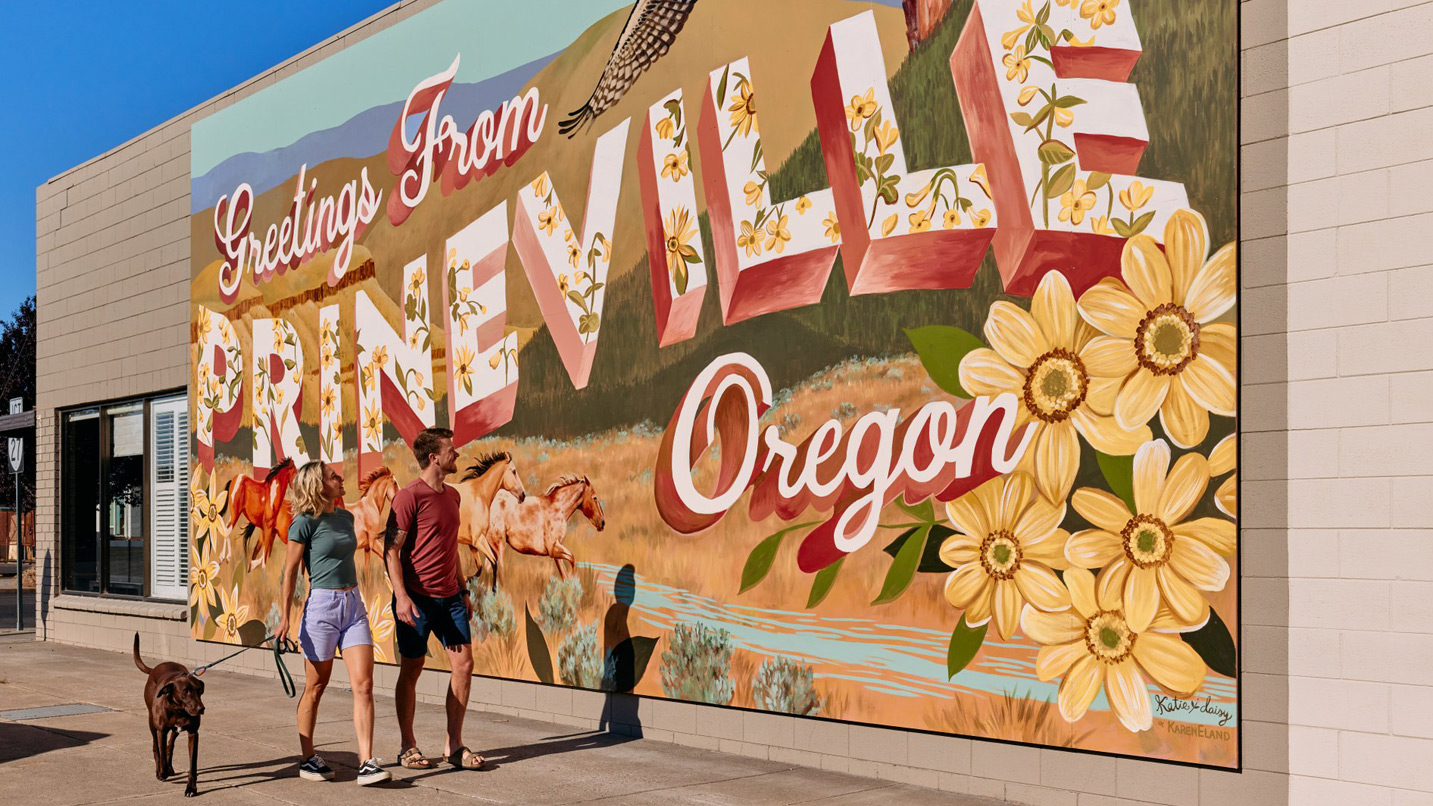 A couple and their dog walk past a "Greetings from Prineville" mural on the outside of a building.