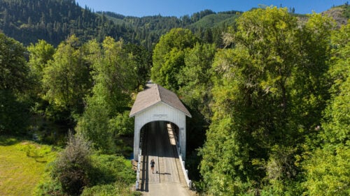 Aerial shot of a white covered bridge surrounded by large canopies of trees. A woman is running on the bridge.