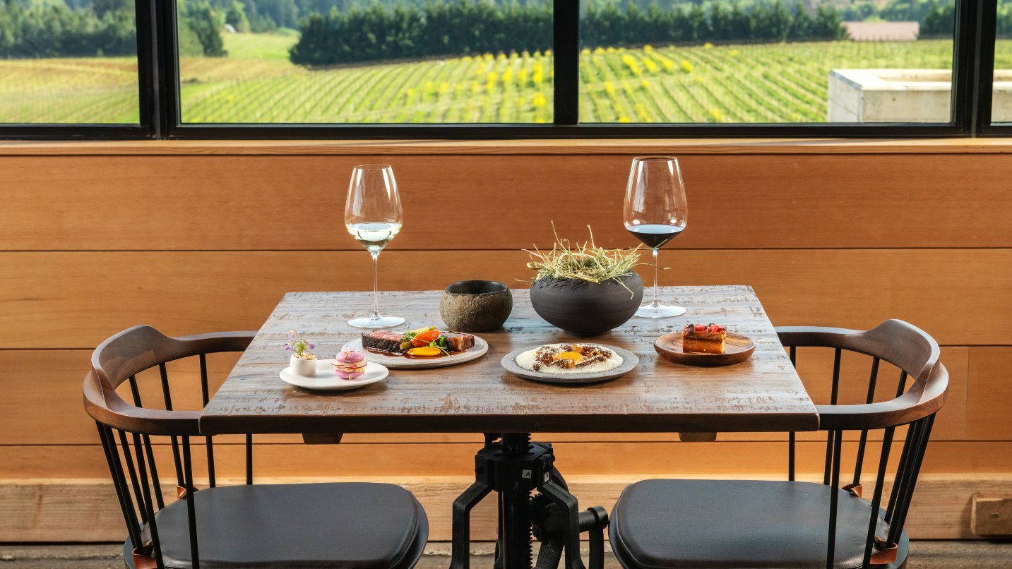 A wooden table holds various plated dishes and two wine glasses, set in a restaurant with large windows overlooking a vibrant vineyard landscape.