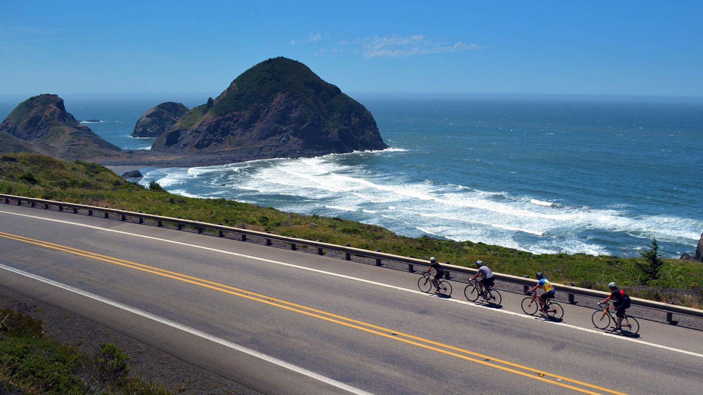 Four cyclists riding on the shoulder of a highway along the coastline with scenery of the waves and rocky formations.