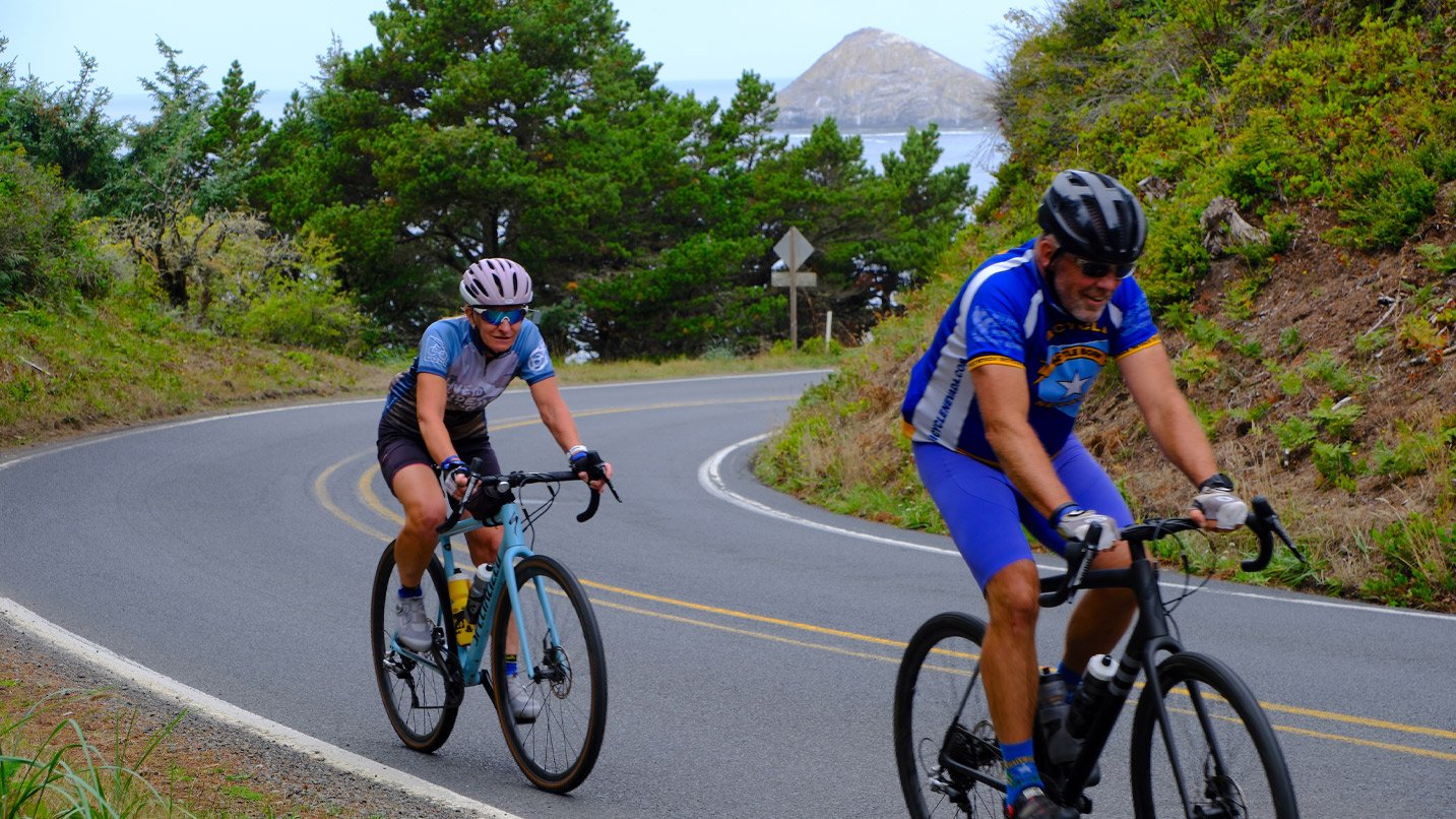 Two cyclists with biking gear round a corner on a highway along the coastline.
