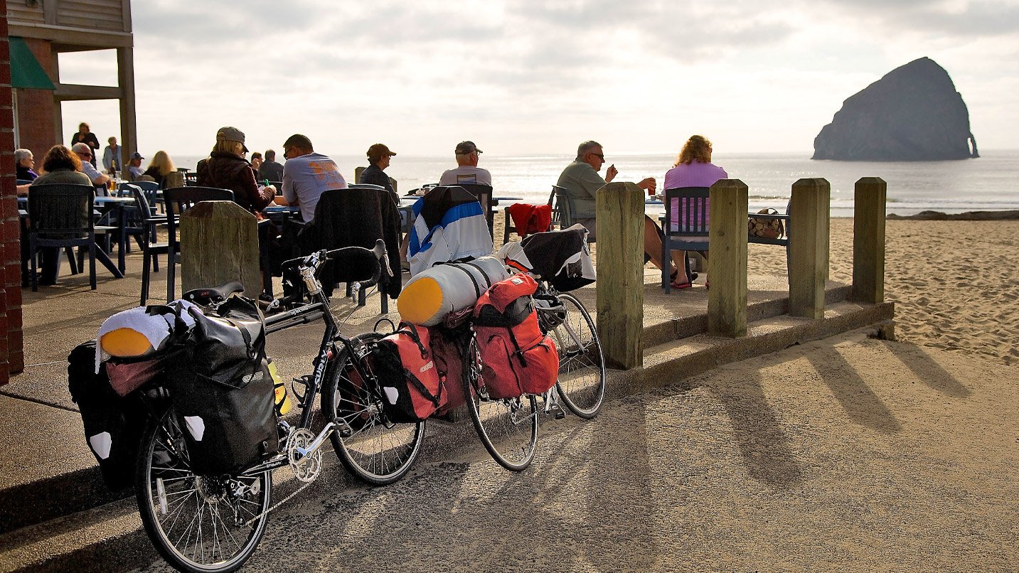 Bikes loaded with gear lean against the wooden posts of a restaurant's outdoor patio where people enjoy a meal.