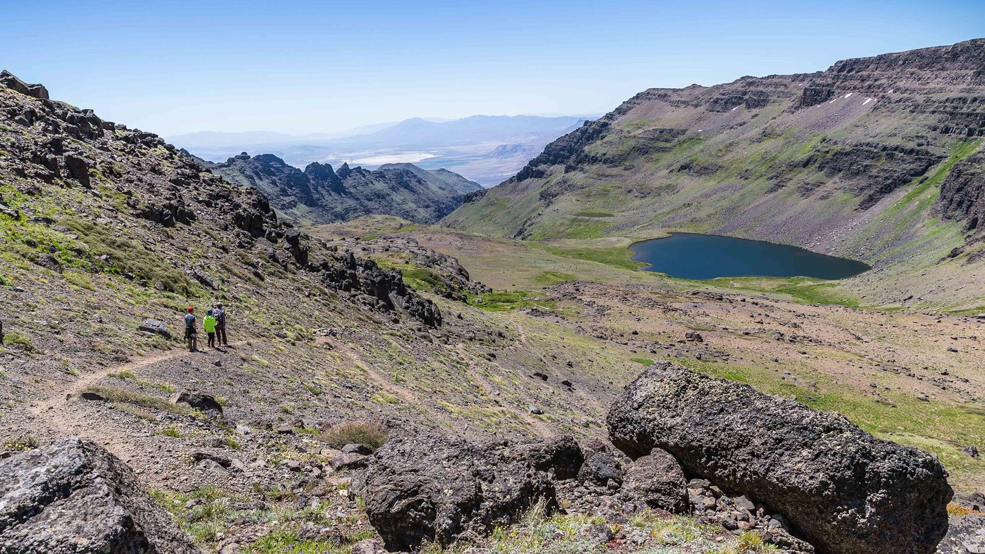 Views of a rocky trail in the Steen Mountain range. 3 People are walking along a marked trail that leads to a small lake.