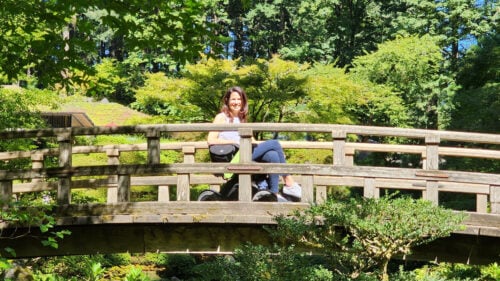 A woman in a power wheelchair on a wooden, Japanese-style bridge surrounded by green foliage.