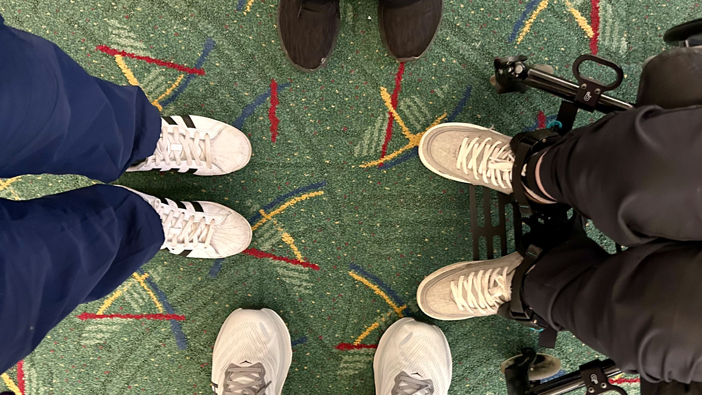 A group of four people standing on the PDX Airport carpet, with their shoes visible and one person in a wheelchair.