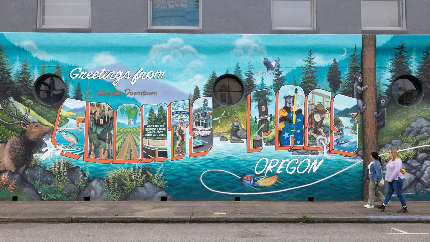 Two women looking at a painted mural of the scenery and experiences of Grants Pass in the vintage postcard style. It reads "Greetings from historic downtown Grants Pass Oregon"