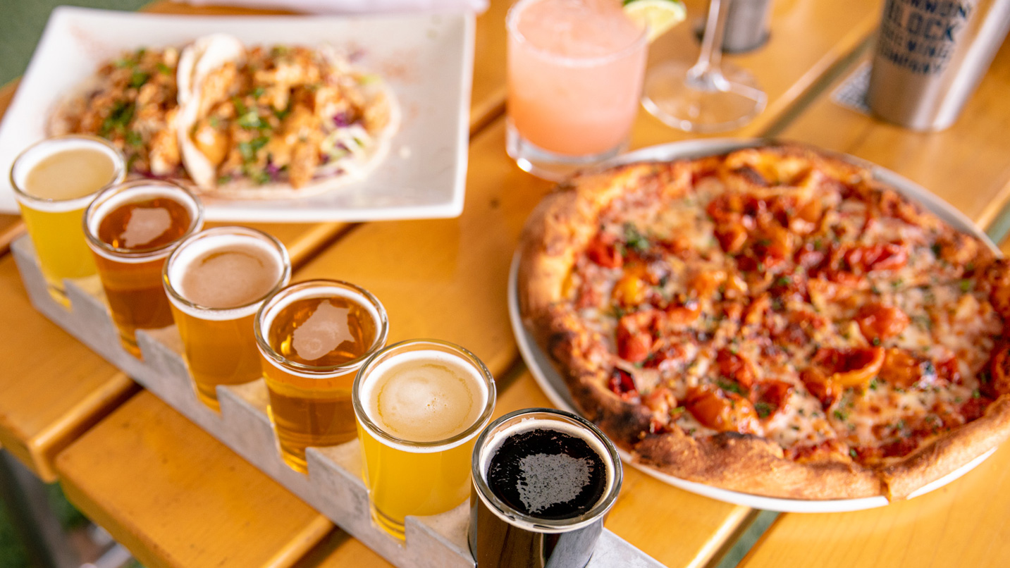 A flight of beer, pizza, tacos and a cocktail on a picnic dining table.