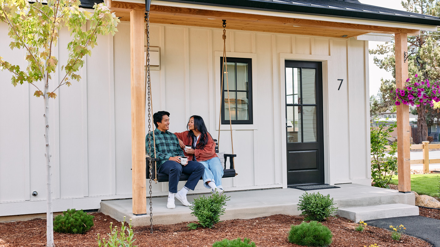 A couple sits on a porch swing in front of a small white cottage.