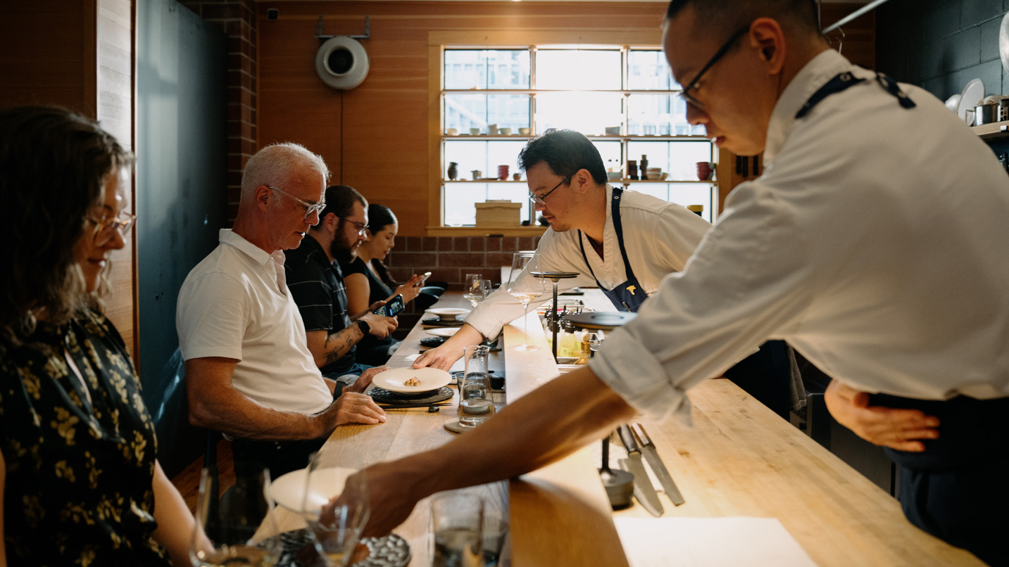 Four people sit at a counter while dishes are being presented in front of them.