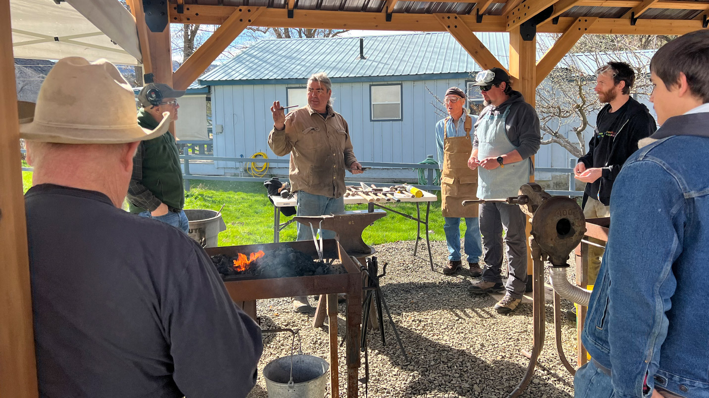 An instructor demonstrates blacksmithing to a small group outdoors.