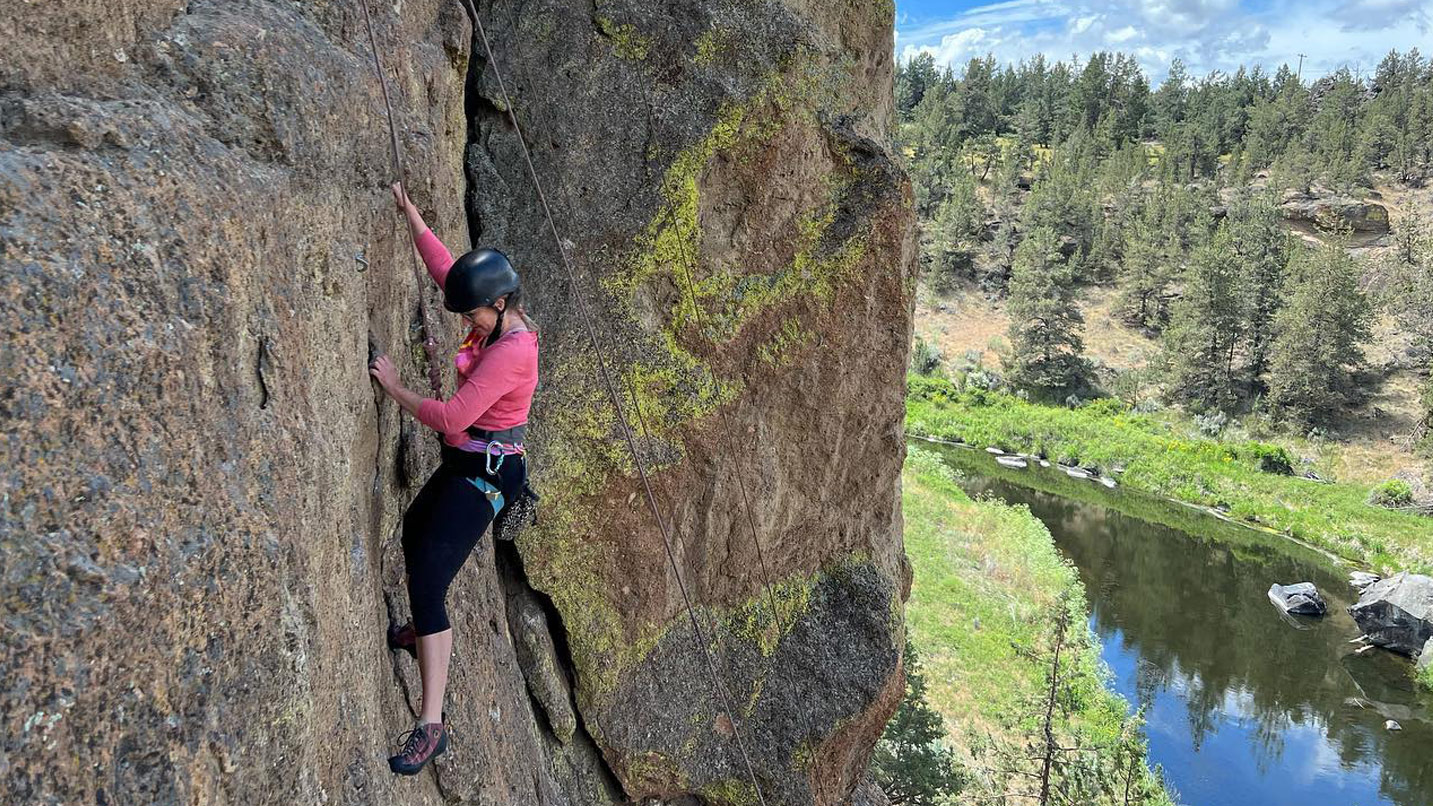 A woman with climbing gear and a harness scaling a cliff.