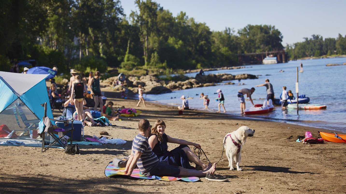 A river bank busy with several people enjoying a sunny day. In the foreground, a couple sit on a beach blanket with their golden dog.