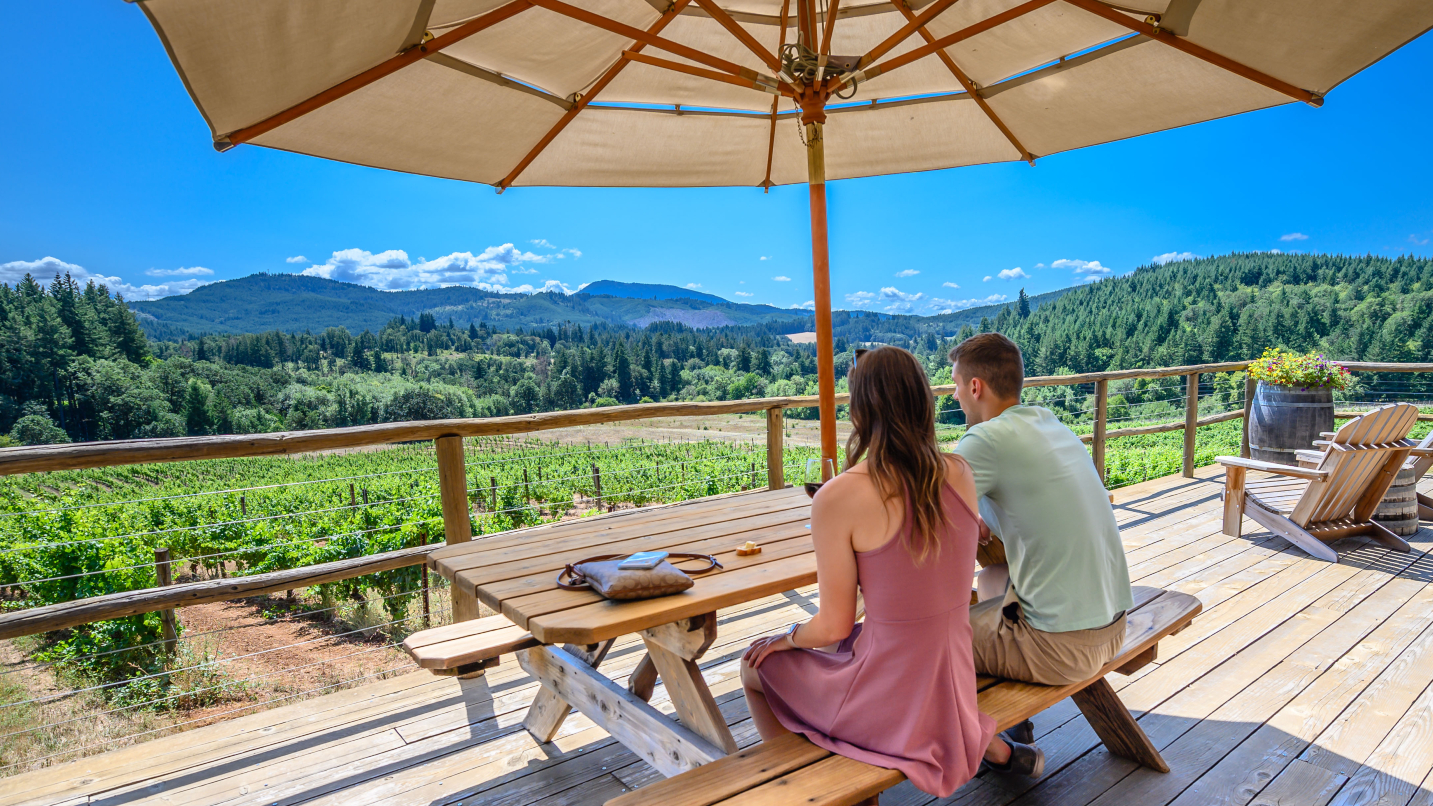 Man and woman sit at patio table on wooden deck with umbrella overlooking vineyard and scenic vista