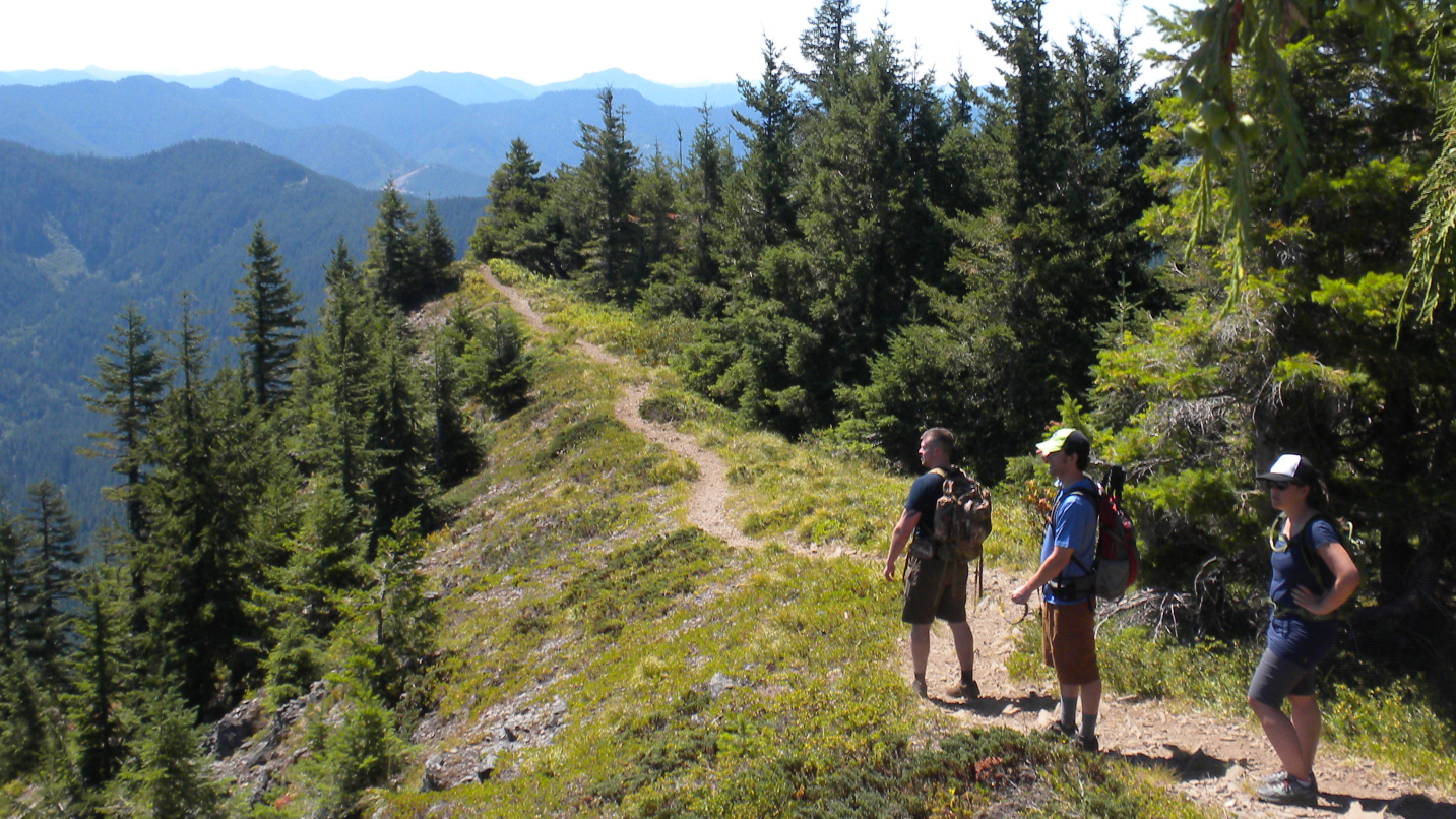 Three people with backpacks stand on a mountain trail looking at a view