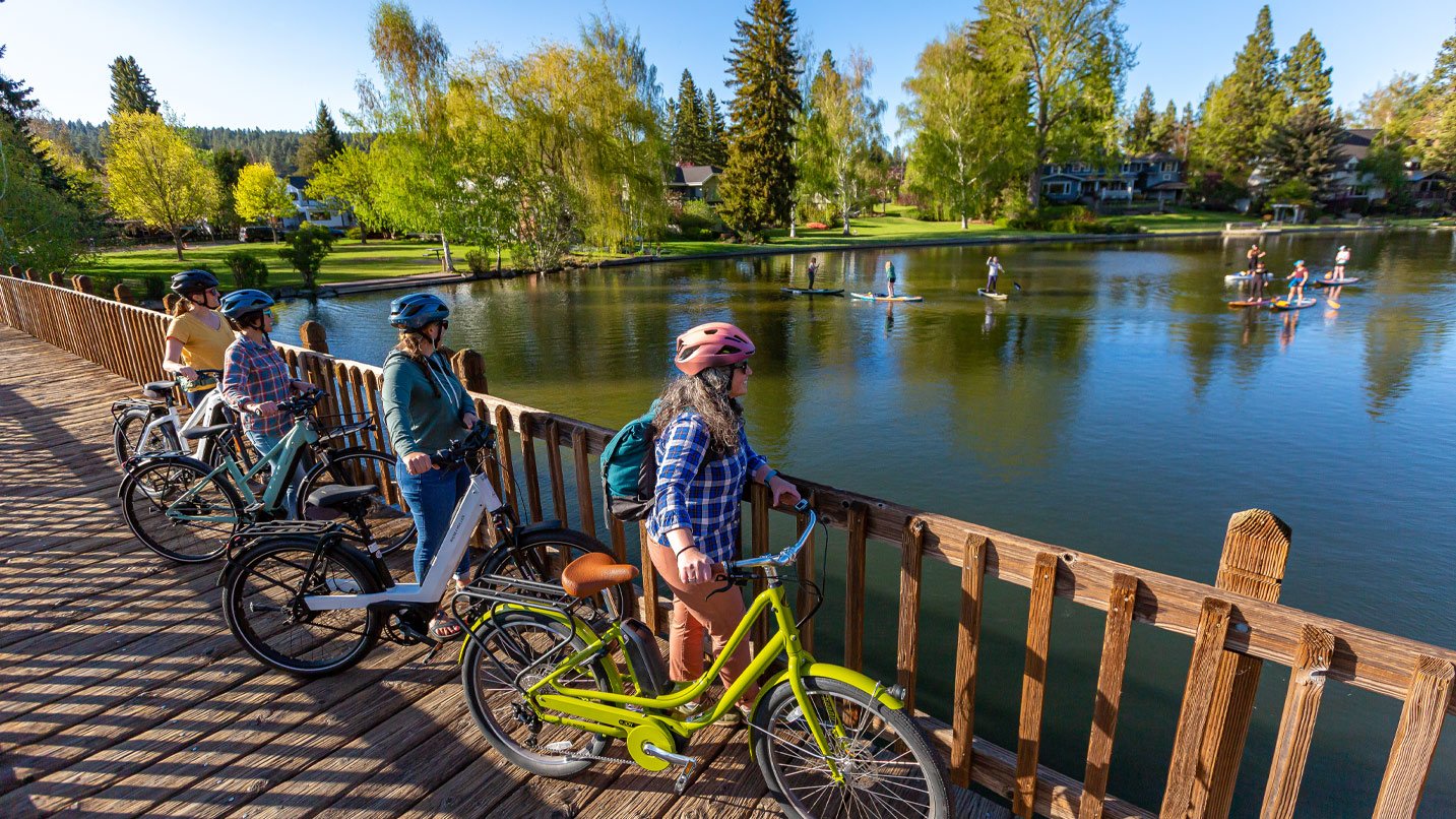 A group of four women stopped on a bridge with their electric bikes looking out at paddleboarders on the Deshutes River.