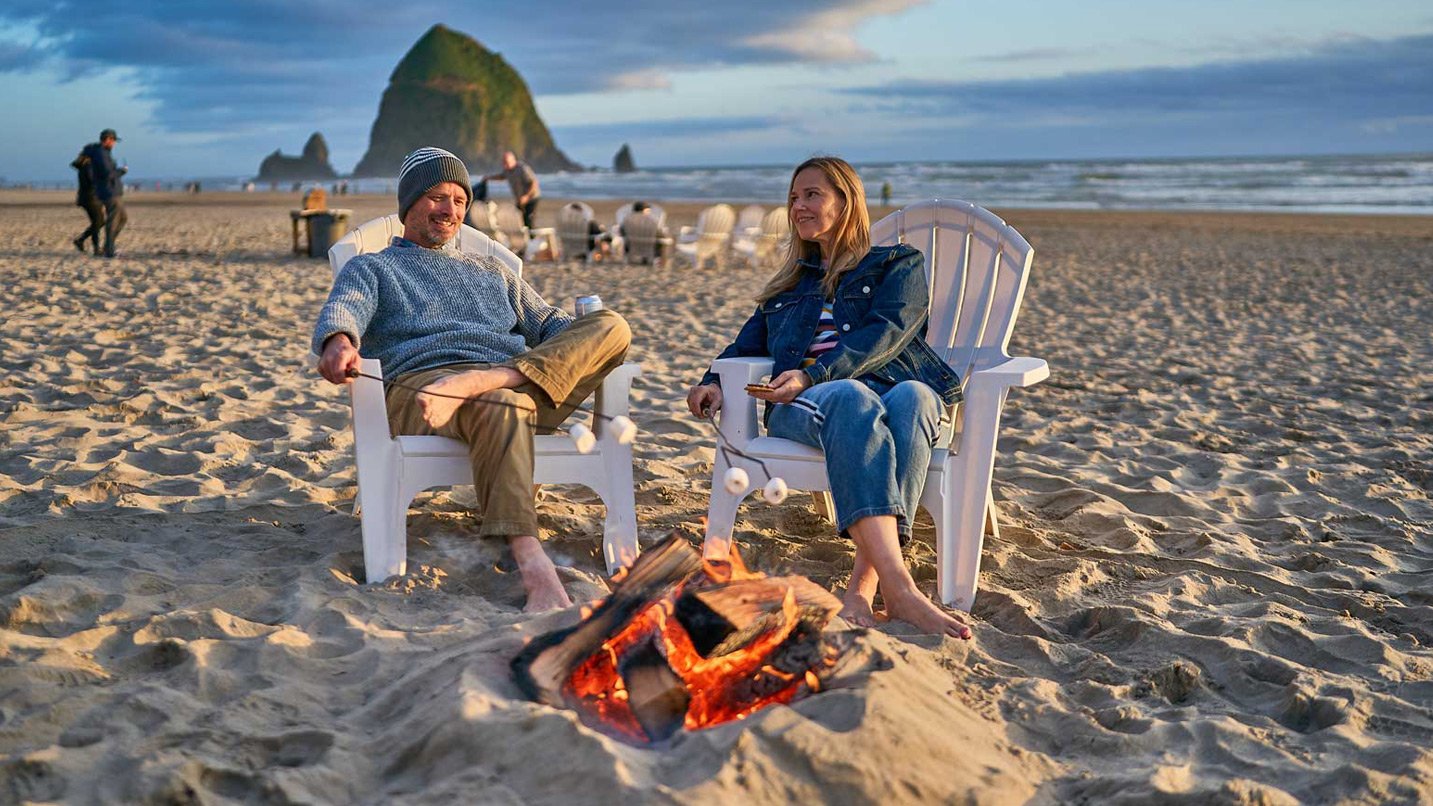 A couple roasts marshmallows outside on the beach.