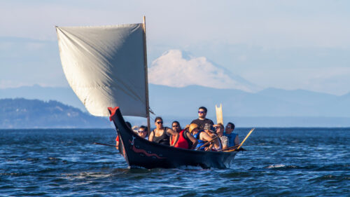 Native Americans paddling a traditional canoe with a wind sail with a snow-capped mountain in the distance.