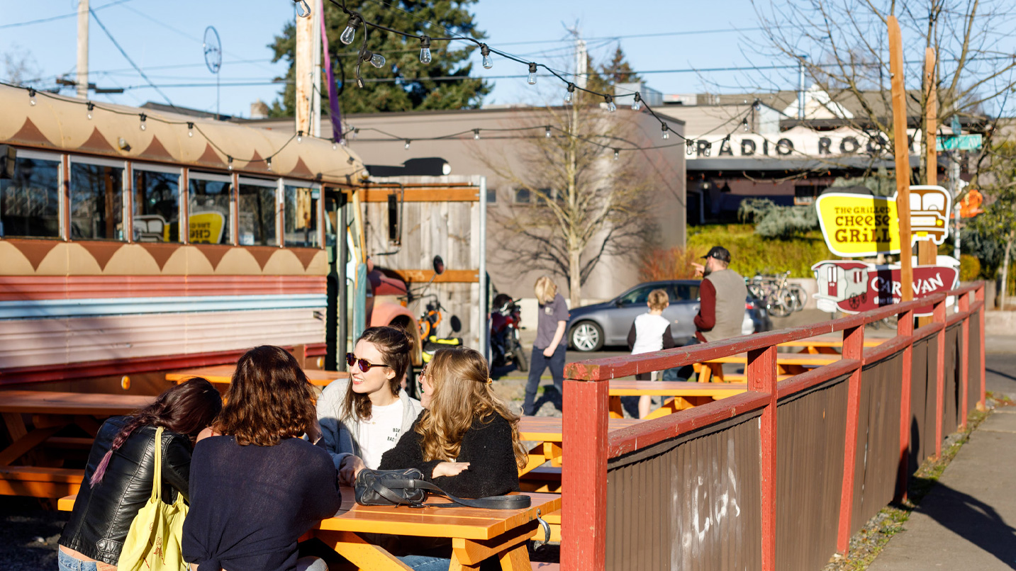 People sit at picnic tables in the sun along a sidewalk with bus in background