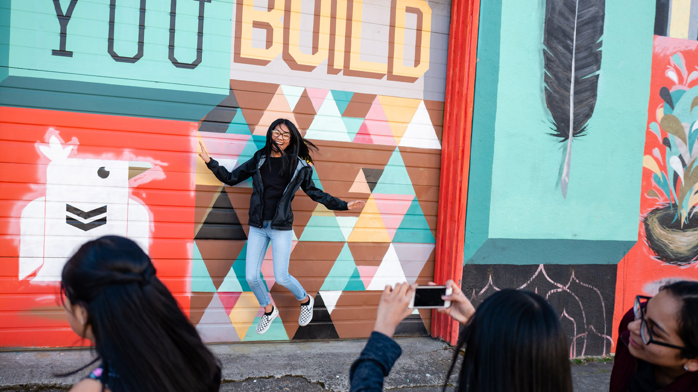 Girl stands in front of a colorful mural and jumps as friends take a photo of her