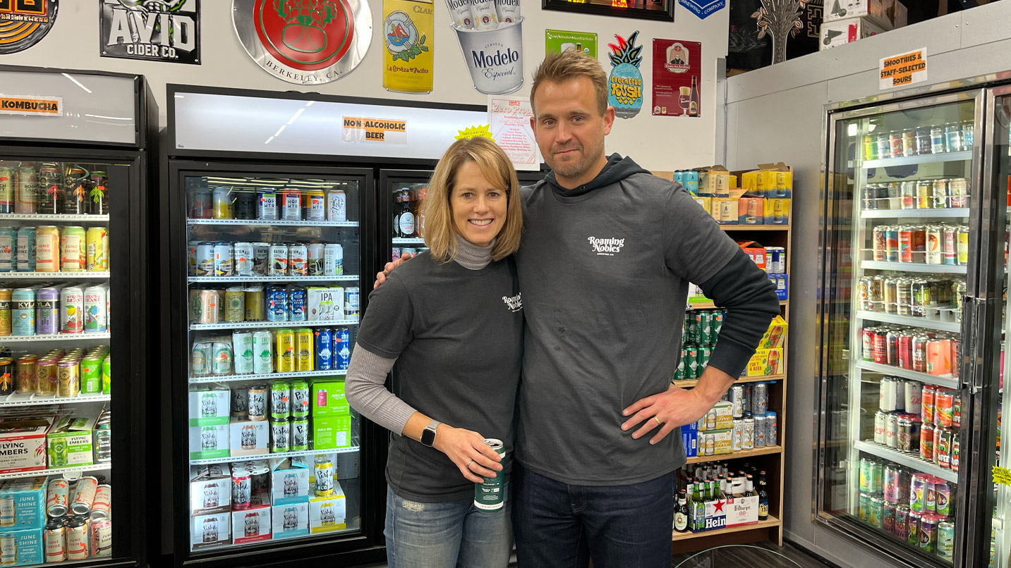 A man and a woman posing in front of store refrigerators filled with alcoholic beverages.