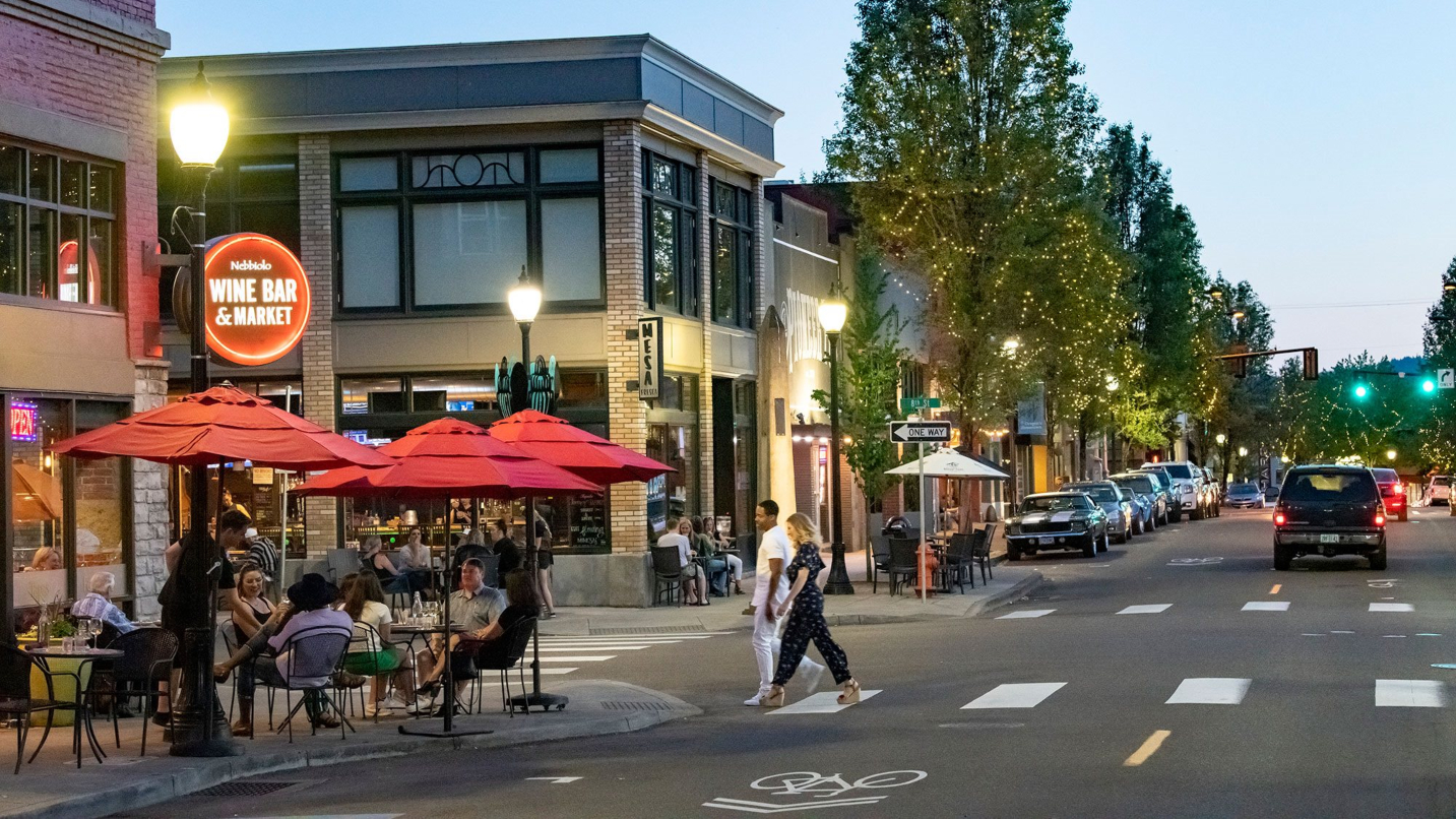 Two people walk across a crosswalk to a wine bar with people sitting on sidewalk under red umbrellas