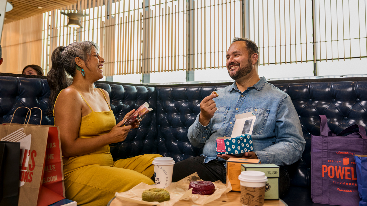 Man and woman sit on black leather couch with snacks and shopping bags