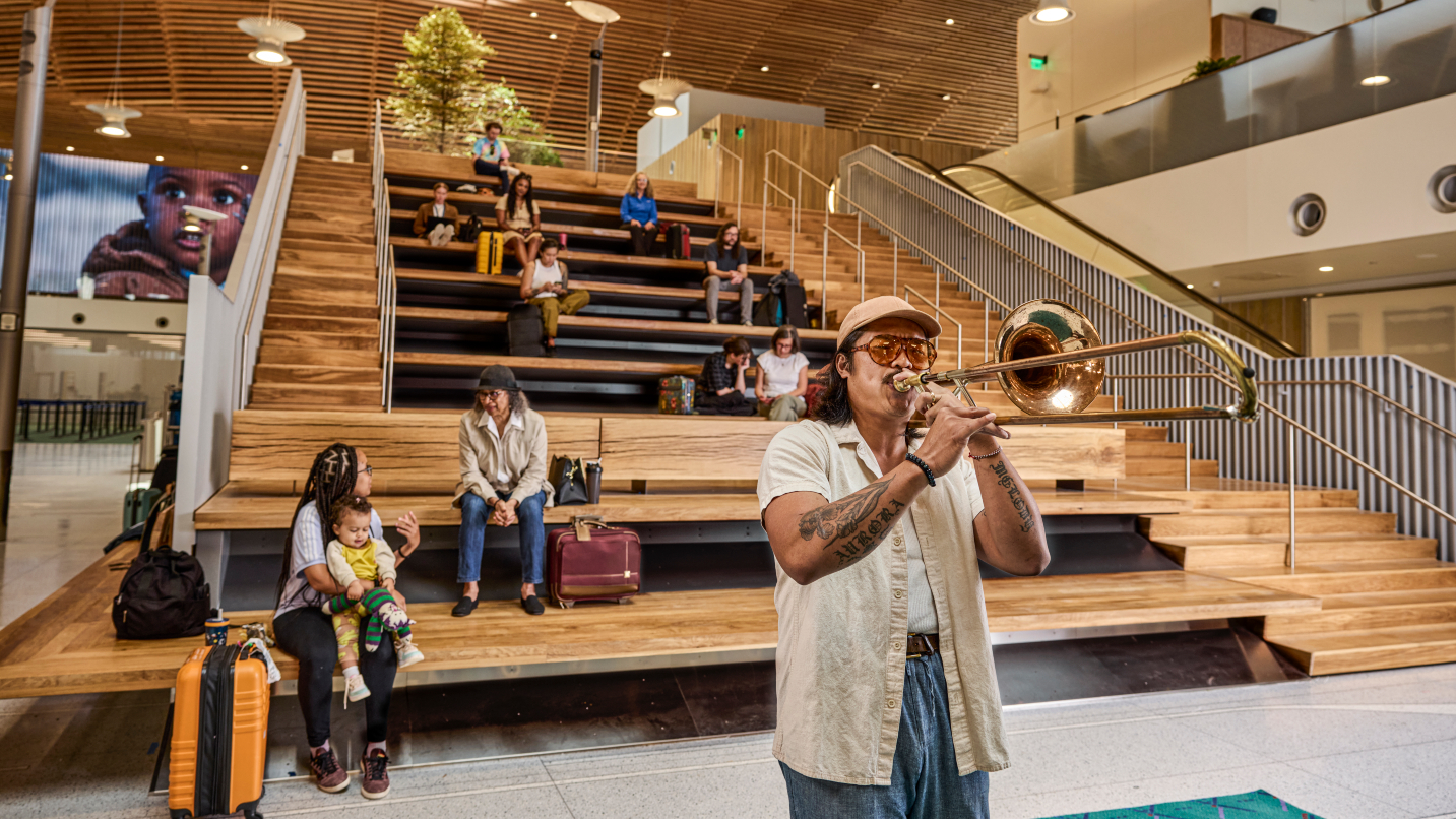 Man plays trumpet as other people sit on wooden bleachers under wood-beam roof