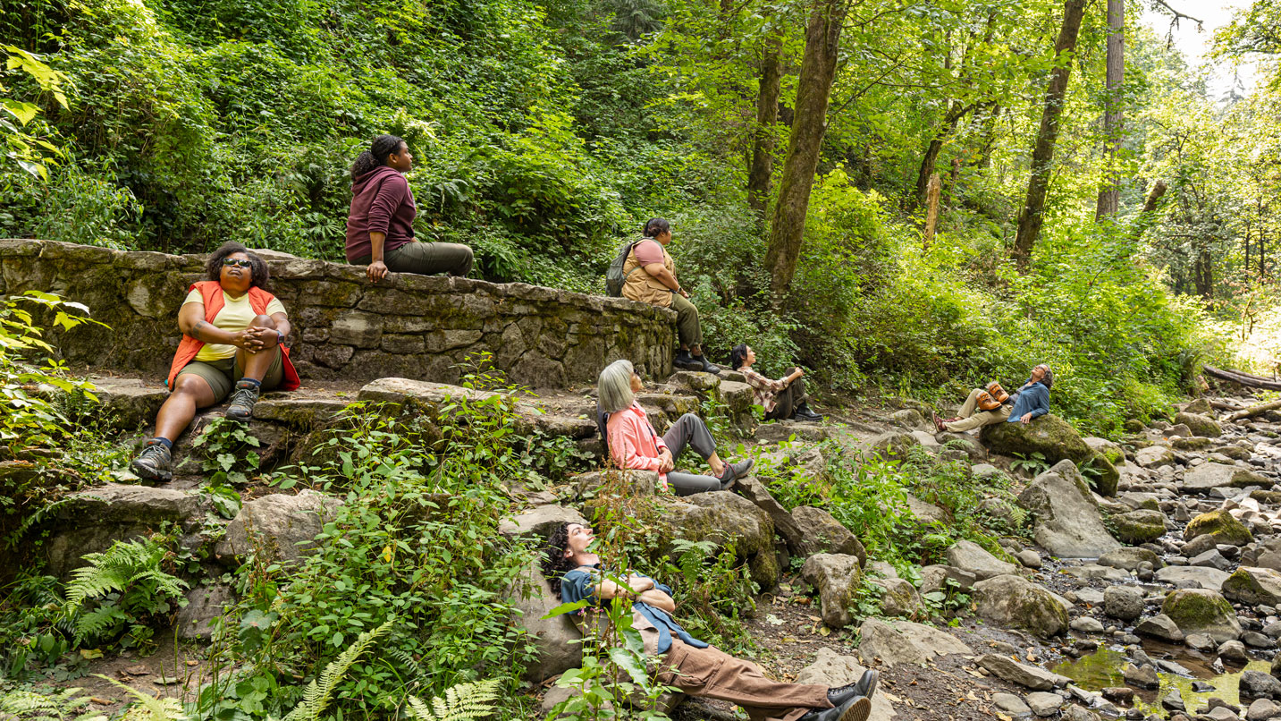 People sit on rocks with trees and creek in background
