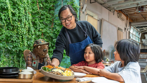 Man smiles while putting platter of food on table with two children and beaver puppet
