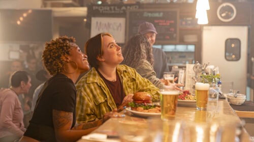 Two women sit at a bar with beer and food in front of them and smile as they look up