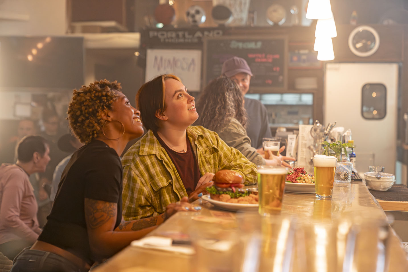 Two women sit at a bar with beer and food in front of them and smile as they look up
